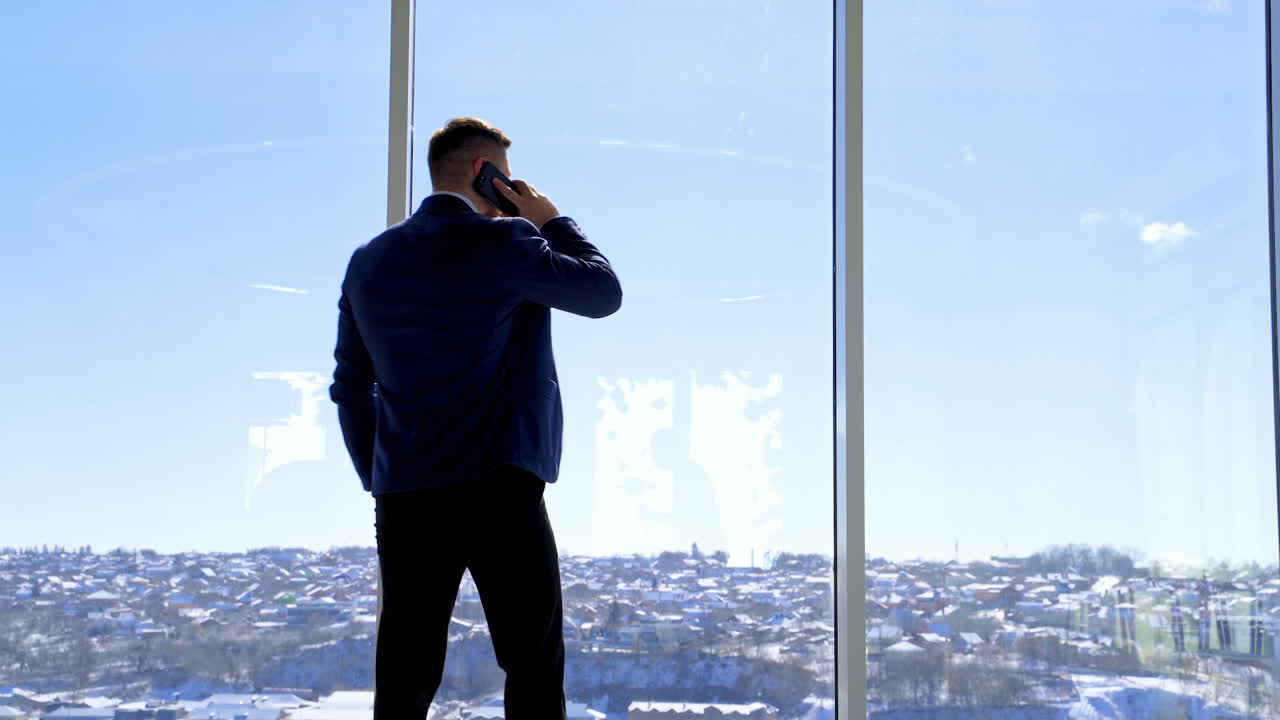 Businessman standing in his office. Rear view of a man wearing a suit talking the phone and looking out of the window. Panoramic window view with a city background.