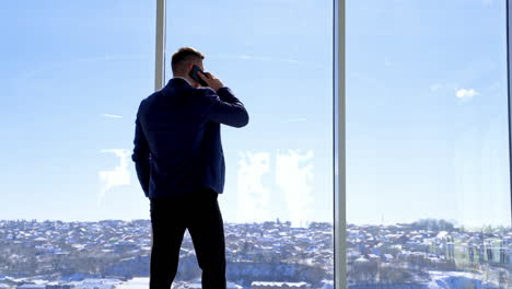 businessman standing in his office. rear view of a man wearing a suit talking the phone and looking out of the window. panoramic window view with a city background.