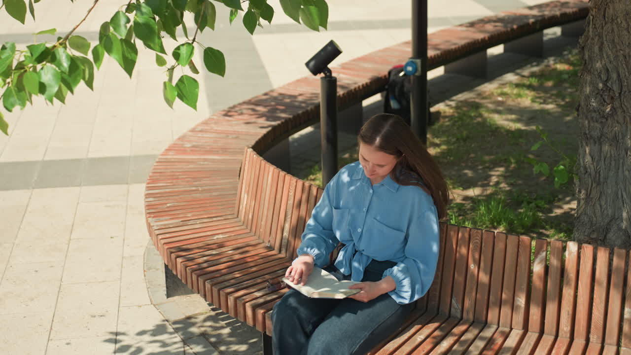 굽은 벽돌 벤치에 앉아 책을 읽고 있는 백인 여성의 오버헤드 뷰, 잎이 무성한 프레임과 기하학적인 포장도로가 있는 공중 공원 구도, 조용하고 한적한 독서 시간, 데님 의상과 집중한 자세, 도시 디자인