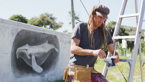 Videoporträt-Eines-Lächelnden-Kaukasischen-Männlichen-Künstlers-Mit-Dreadlocks,-Der-Die-Farbe-Eines-Walwandgemäldes-An-Der-Wand-Hält