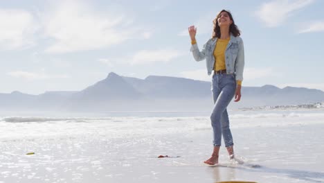 Happy-mixed-race-woman-walking-barefoot-on-the-beach-by-the-sea,-enjoying-the-view-and-smiling