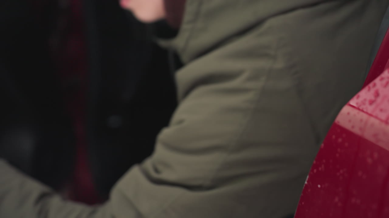 Close up of red car as man exits vehicle during light snowfall, water droplets visible across exterior door, with person in winter jacket moving from driver seat into snowy environment