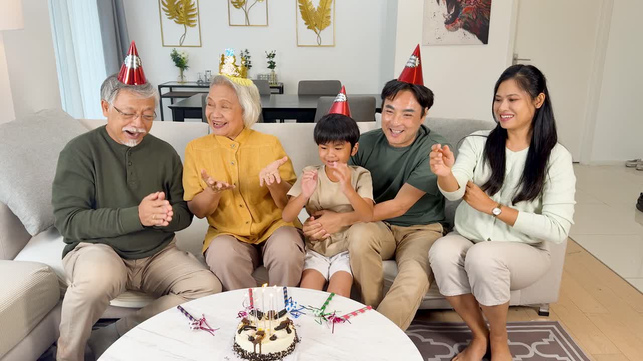Four-generation Asian family claps and smiles as child blows out birthday cake candles indoors