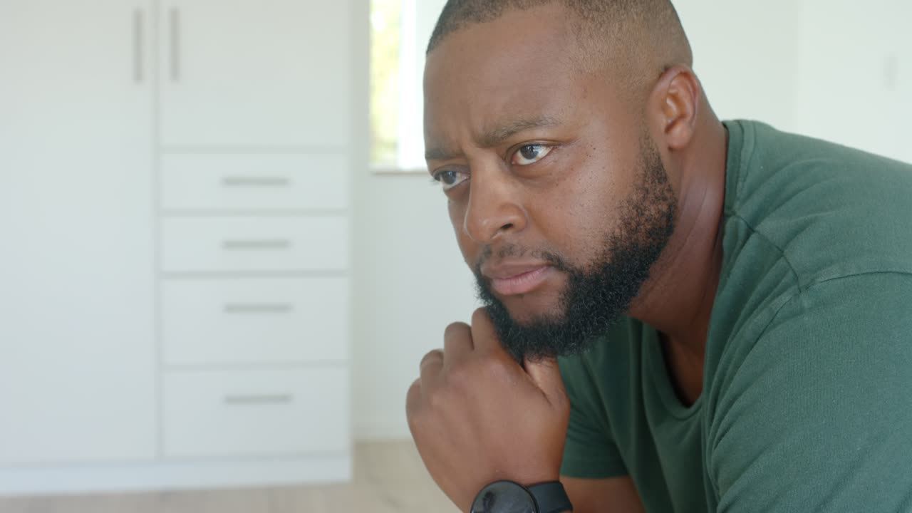 African male sitting in bright room leaning shifting gaze reacting to camera moving wearing tee