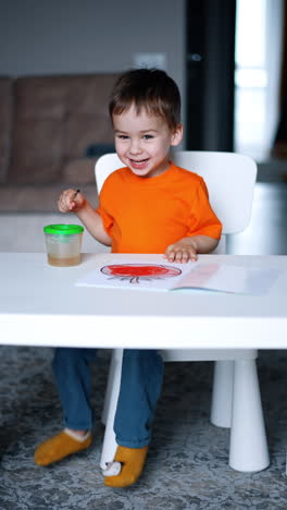 cheerful kid sitting at the desk stepping his feet joyfully. happy smiling toddler painting with a brush. vertical video.