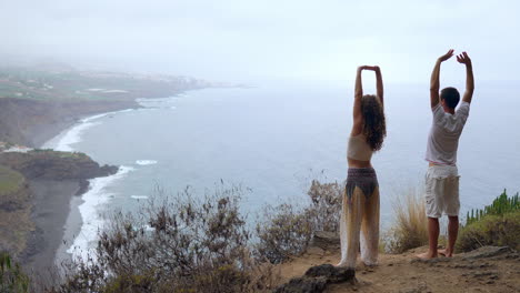 atop a cliff, a man and woman lift their hands, inhaling the ocean breeze as they engage in yoga, embracing the calming atmosphere