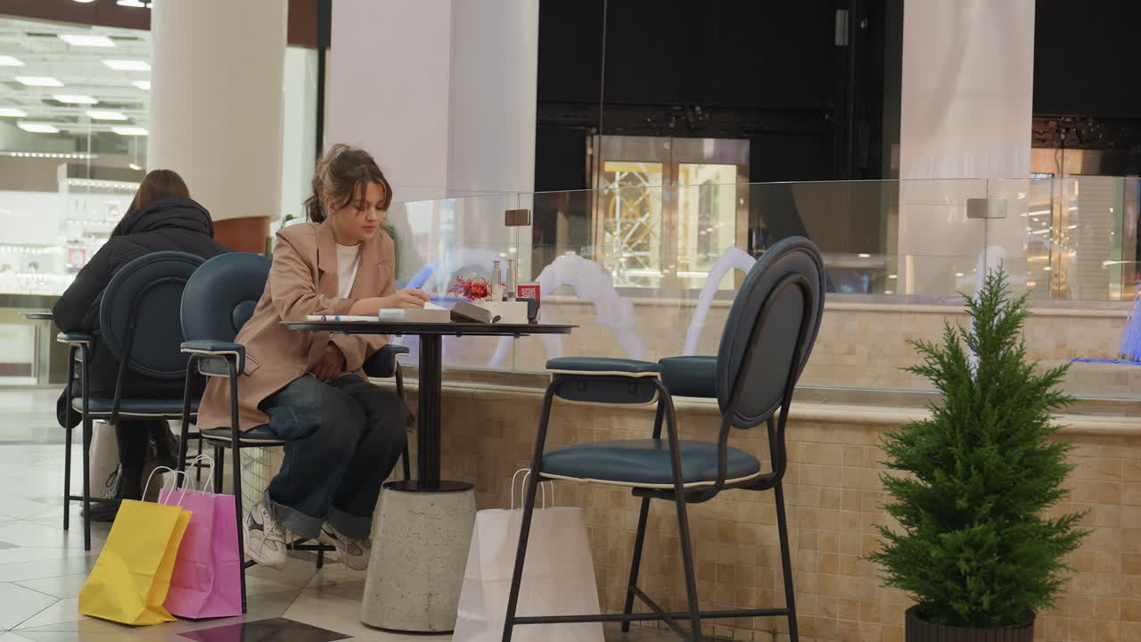 Una mujer relajada toma notas cerca de unas bolsas de la compra. Una compradora informal escribe una nota durante una pausa a mediodía en una cafetería. Una mujer con ropa informal disfruta de un café mientras toma notas al lado de sus bolsas de la compra.