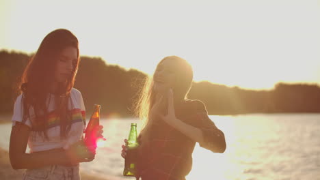 two female students with perfect young bodies are dancing in short t-shirts with beer on the beach party at sunset. their long hair is flying on the wind.