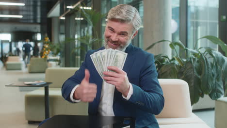 mature businessman counting dollar banknotes showing thumbs up and looking at camera in office lobby