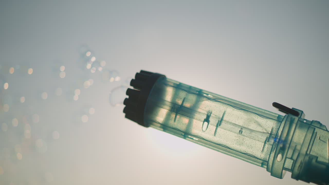 A plastic bubble gun releasing a stream of colorful bubbles under a bright, sunny sky. The bubbles capture and reflect the sunlight, creating a playful and cheerful atmosphere