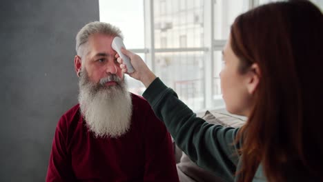 over the shoulder a brunette girl in a green sweater measures the temperature with an electronic thermometer of her elderly father with gray hair and a lush beard in a red t-shirt while sitting on the sofa in a modern room