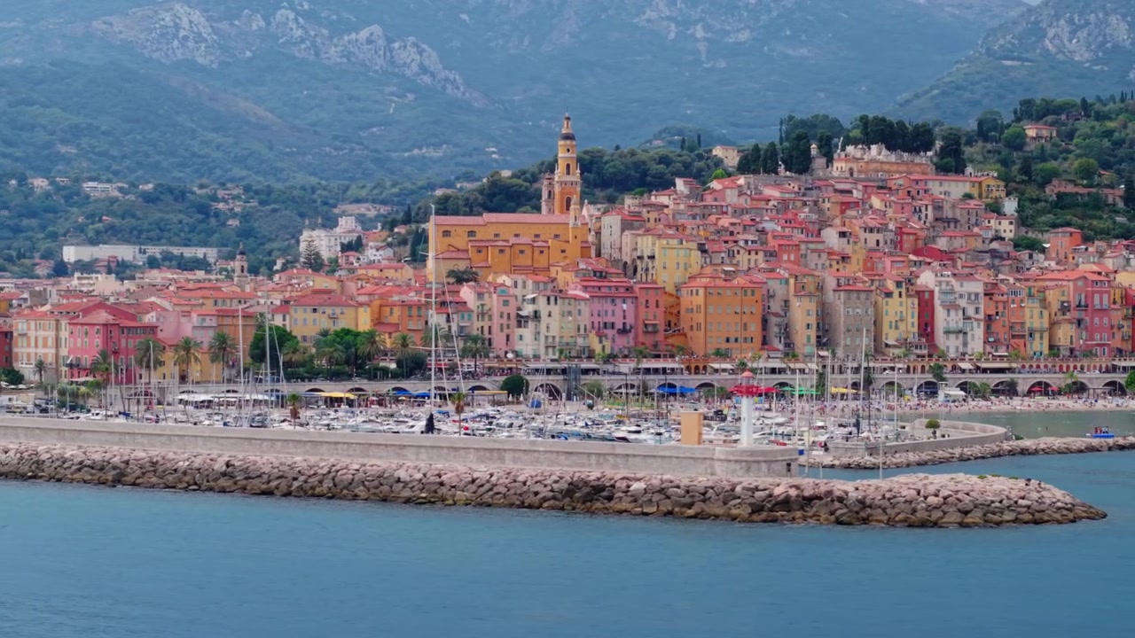 Case colorate e chiesa di Mentone si affacciano sul porto turistico e sul mare con montagne sullo sfondo