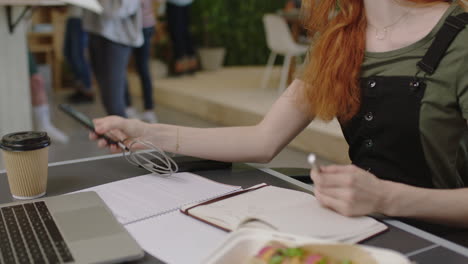 young caucasian business woman writing notes enjoying study listening to music wearing headphones female student using smartphone checking messages in diverse office workplace