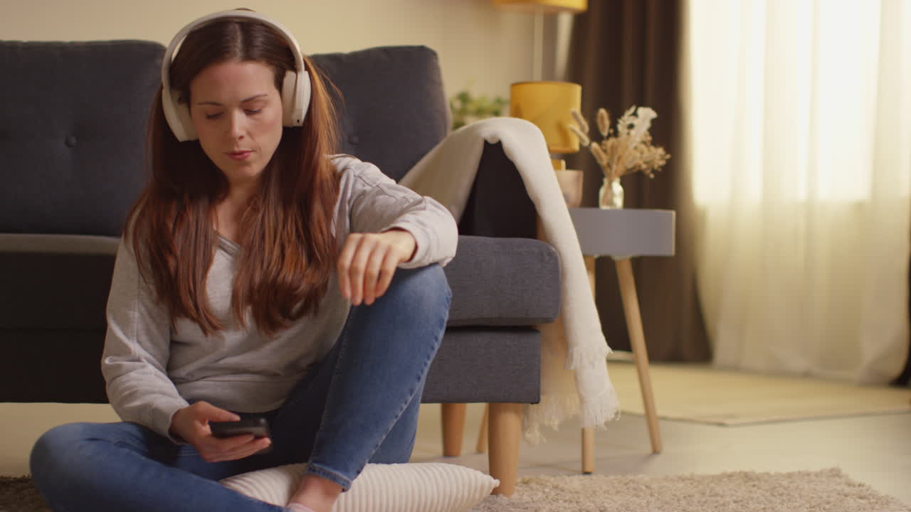 Woman Wearing Wireless Headphones Sitting On Floor At Home Streaming Music From Mobile Phone 13