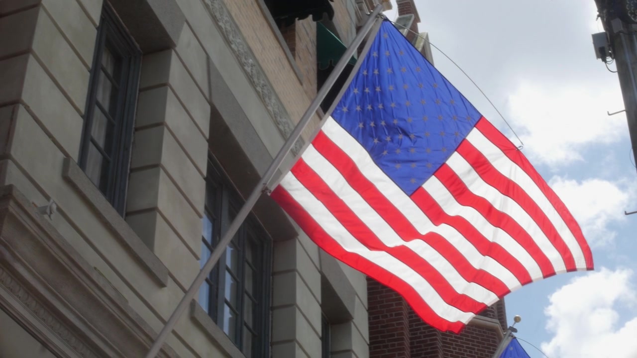 Bandiera che sventola da un edificio del centro contro un cielo nuvoloso e luminoso di giorno. Primo piano dal basso che mostra le strisce americane accanto a dettagli architettonici urbani in mattoni.