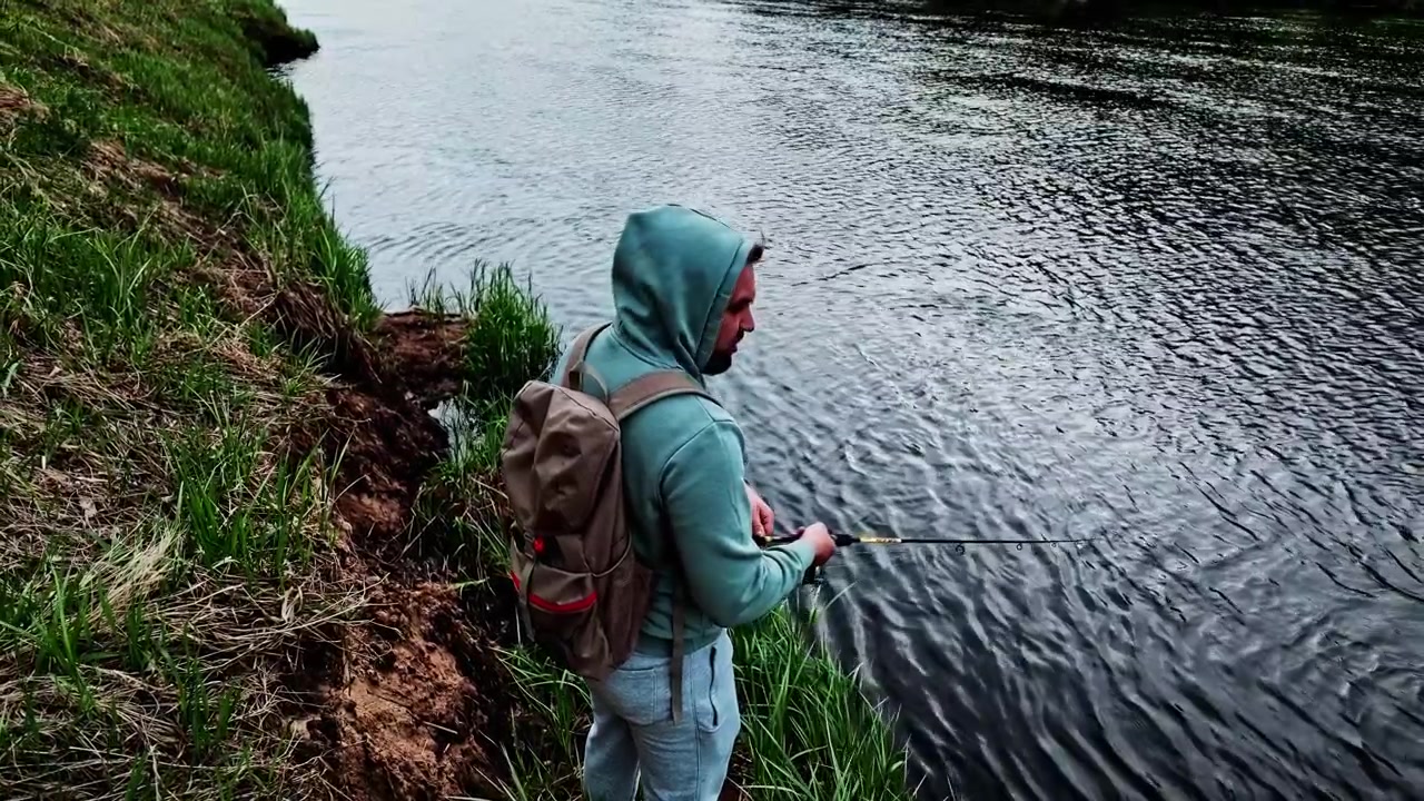 Nelayan berjaket hoodie memegang joran pancing putar dan memancing dari tepi sungai berumput