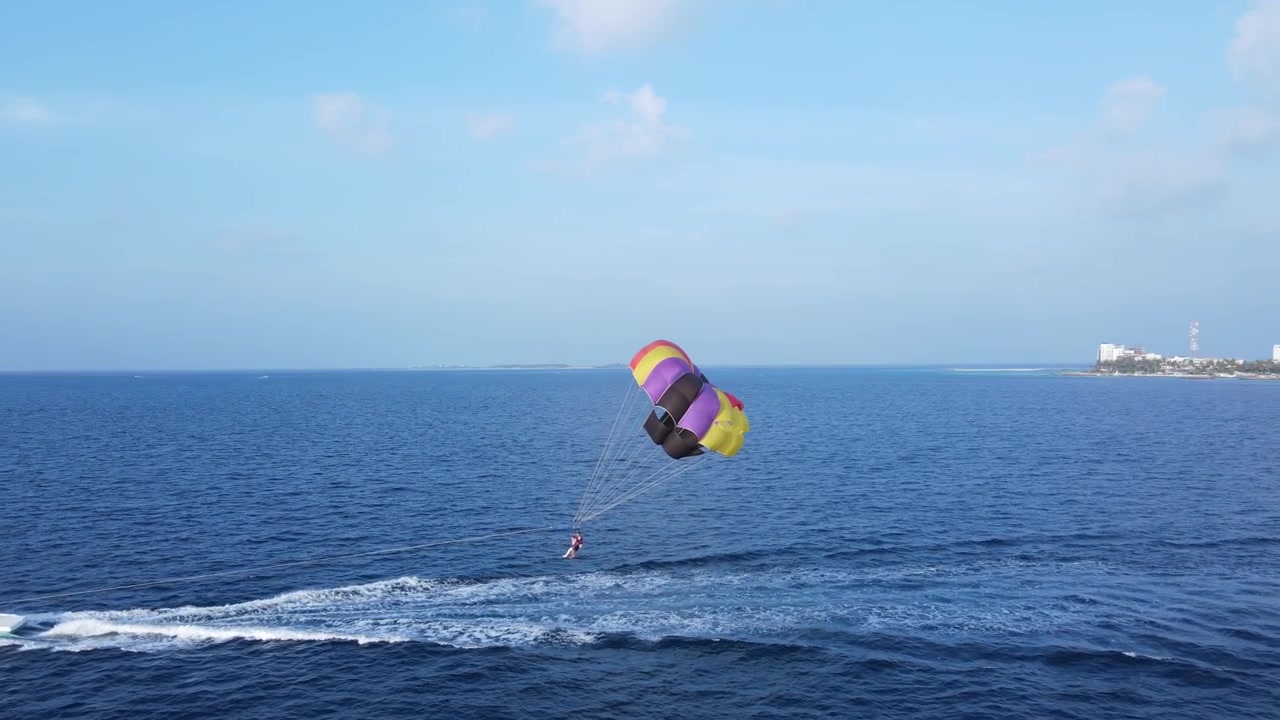 Drone view of couple people parasailing with a colorful parachute pulled by a speedboat over blue tropical ocean. The scene highlights adventure, summer vacation, and water sports.