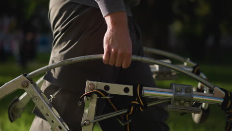 close-up of human hand gripping metal bar, showing hand, lower arm in grey long-sleeved shirt, dark-grey brown work pants. blurred green grass background suggests outdoor setting