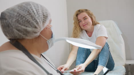 client wearing white top and jeans seated on white chair filing toenails under bright salon light while technician in protective gear sits close, creating a calm and focused personal care setting