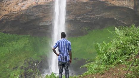 African-youth-standing-at-the-base-of-a-large-and-powerful-waterfall-in-tropical-Africa