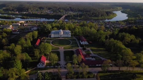 drone footage circling a historic mansion surrounded by lush greenery in a picturesque riverside town. the sweeping aerial view captures nearby buildings trees and a bridge over the river at sunset.