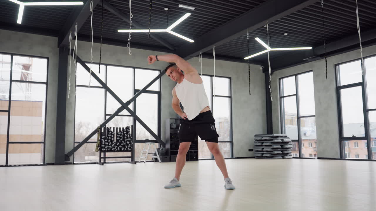 Fitness trainer performing side stretch in spacious gym room with large windows, wearing white tank top and black shorts, equipment racks visible, conveying warm up focus and muscle activation