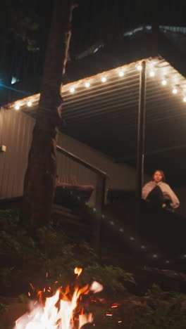 mujer solitaria se sienta en un sillón en la terraza. patio de la casa de glamping con hoguera ardiendo en la noche de verano. relajación en el edificio rodeado de bosque