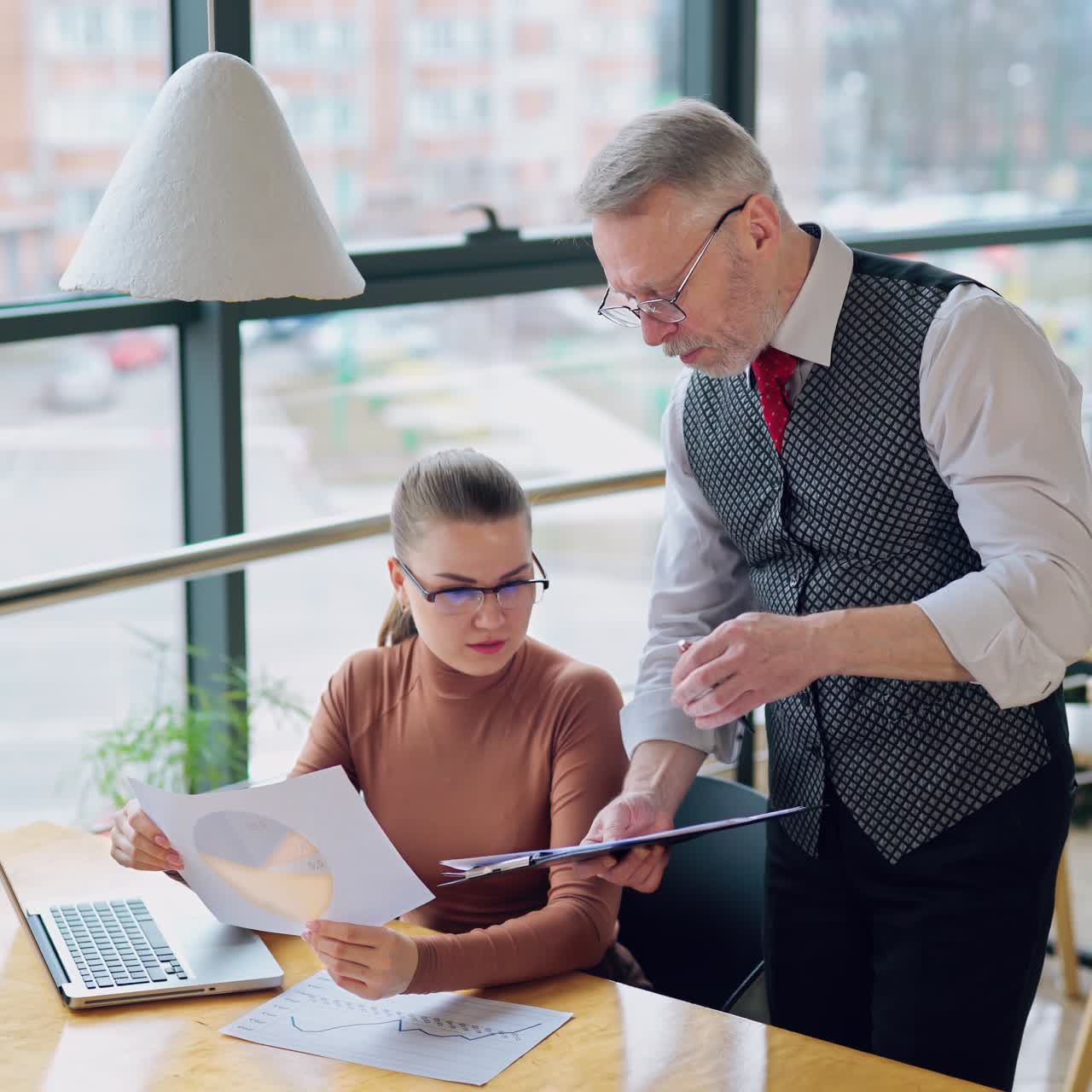 Two colleagues discussing the work in the company. Mature businessman talking to his secretary near window on the city background.