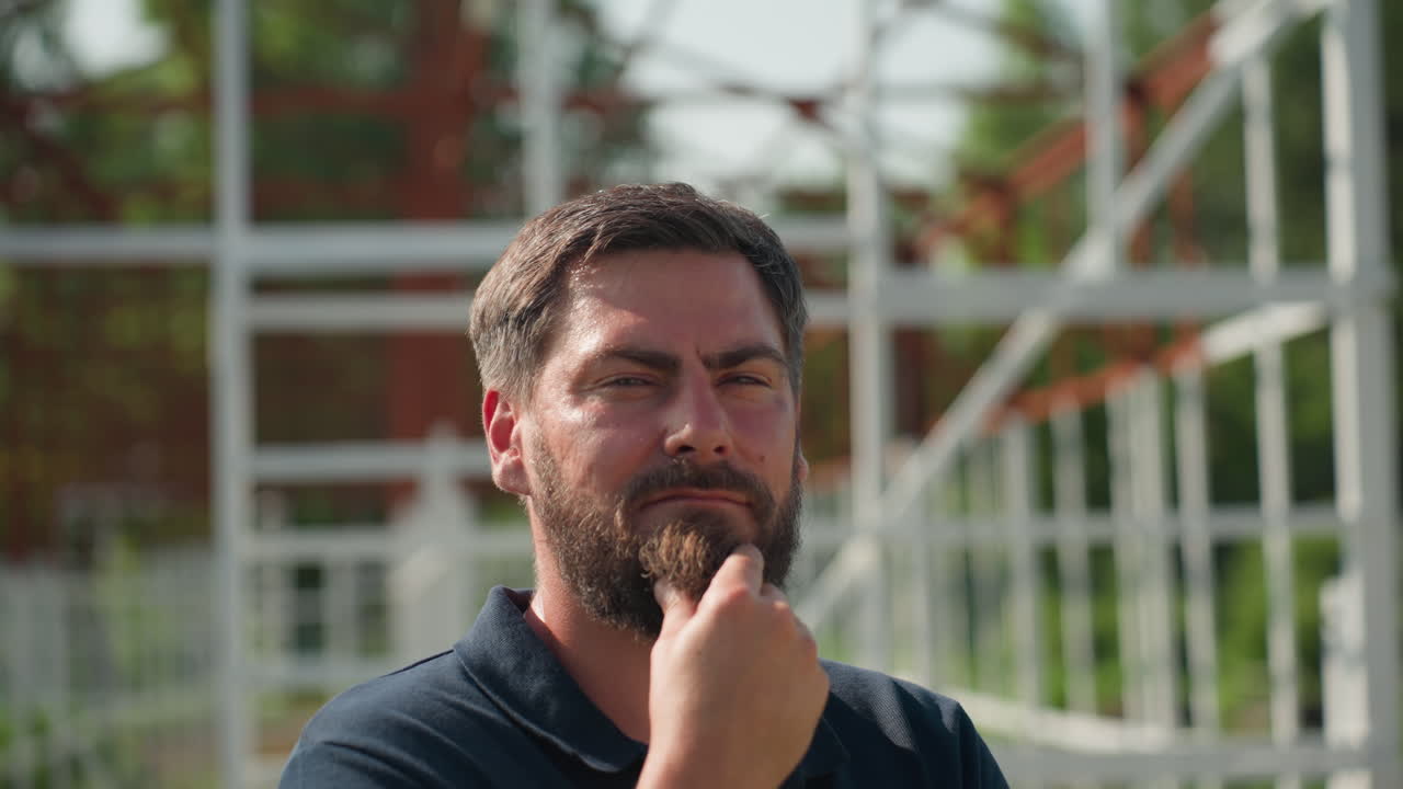 young man close up touching beard while thinking beside metal frame structure outdoors summer light soft shadows on face expression contemplative serious planning future project
