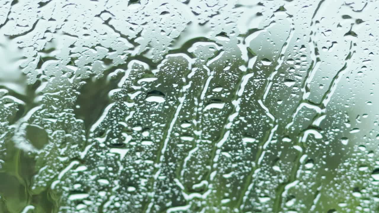Close-up view of raindrops trickling down a glass surface with a blurred green background.