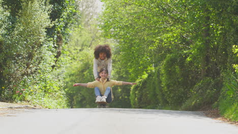 children having fun with boy pushing girl on skateboard on country road