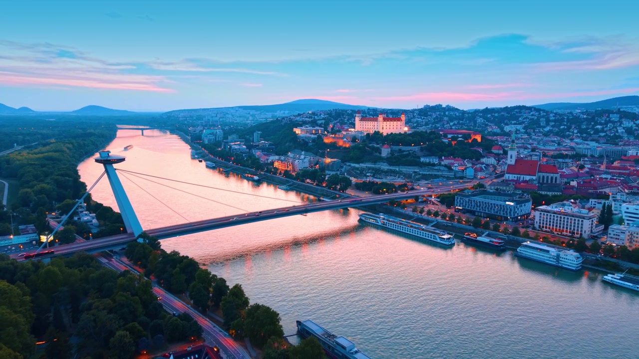 Luftperspektiv af Most SNP-broen og Bratislava Slot ved solnedgang. Skråstagsbro-konstruktion og oplyst højborg reflekteret i floden under skumring i Slovakiet.