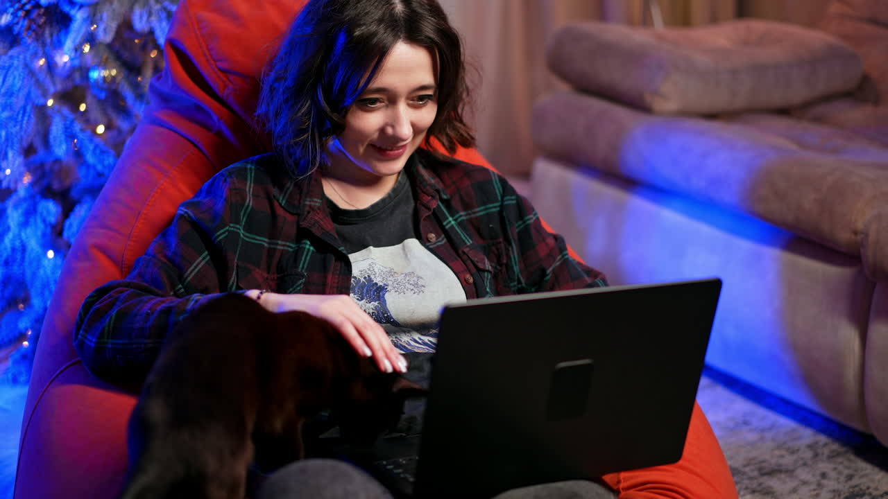 Happy relaxed Caucasian lady sits in armchair holding a laptop. Smiling woman works on computer and pets her black cat. Christmas tree at backdrop.