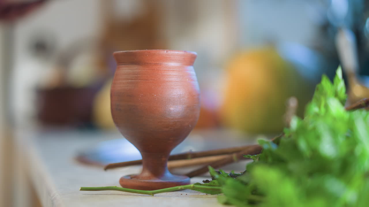 Caneca de barro rústica na bancada, luz suave da manhã, colher de pau e salsa fresca, cozinheiro desfocado ao fundo preparando refeição, atmosfera acolhedora de culinária caseira com profundidade de campo rasa e bokeh suave