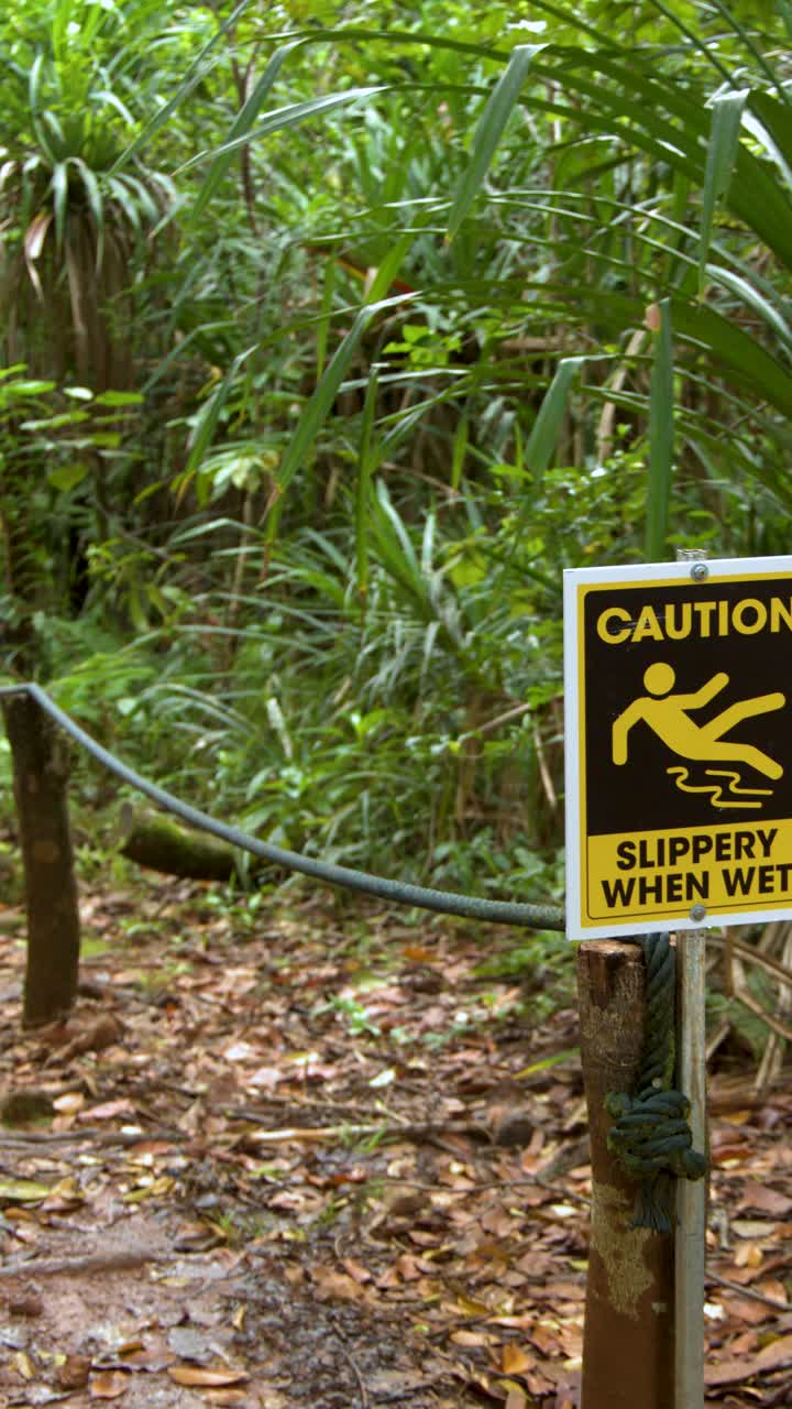 A yellow caution sign warns hikers of slippery conditions on a lush tropical forest path