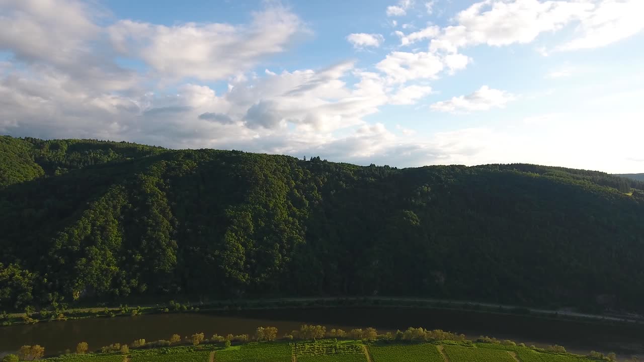 High forward flying drone aerial view of a forest overgrown hill and fields bellow