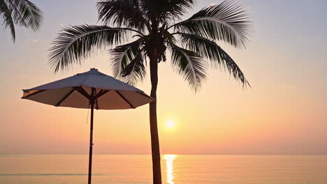 Vista-Perfecta-De-La-Puesta-De-Sol-Roja-Desde-El-Salón-Del-Complejo-Hotelero-Tropical-Cerca-Del-Mar,-Sombrillas-De-Playa-Y-Silueta-De-Palma-De-Coco-En-Primer-Plano