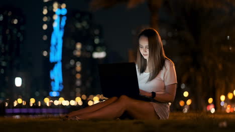 una mujer joven en una noche de verano sentada en la hierba en la ciudad mira la pantalla del portátil y escribe un mensaje en el teclado. programador freelance. trabajo nocturno remoto
