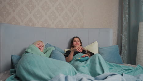 two girls lie in bed under teal blanket, one eating popcorn and laughing, the other resting with content expression, creating cozy atmosphere of friendship, comfort, and playful relaxation in bedroom
