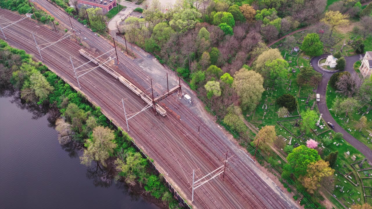 Vista aerea con drone a picco su più linee ferroviarie elettrificate che corrono lungo il fiume Schuylkill e un cimitero verde con fogliame primaverile