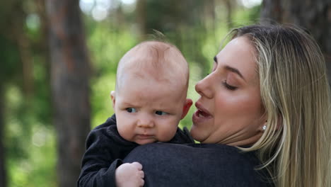 blonde caucasian woman holding her baby. loving mother kissing her child's tiny ear. close up. nature at backdrop.