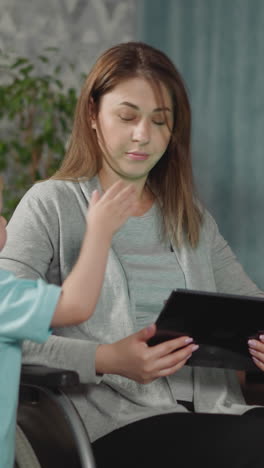 mother listens to blond son talking while watching cartoon on tablet. cute toddler and preschooler girl stand near mom sitting in wheelchair at home