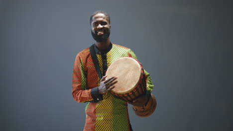 Portrait-of-young-cheerful-african-american-man-in-traditional-clothes-playing-a-drummer-and-smiling-at-camera