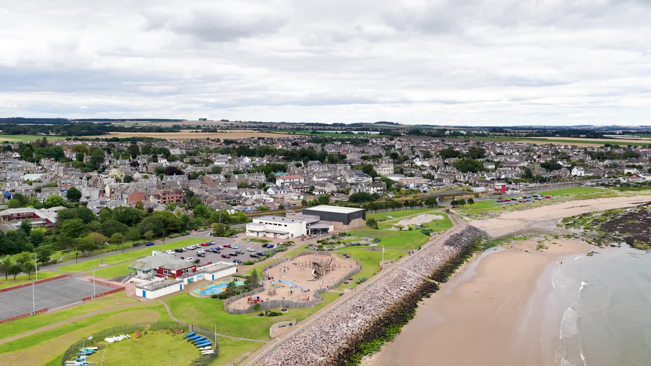Drone glides from urban Dundee over green spaces to sandy coastline under overcast daylight