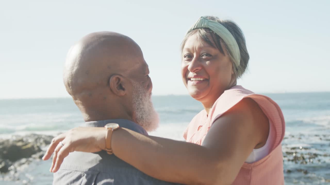 Retrato de casal afro-americano idoso feliz se abraçando no calçadão à beira-mar, em câmera lenta