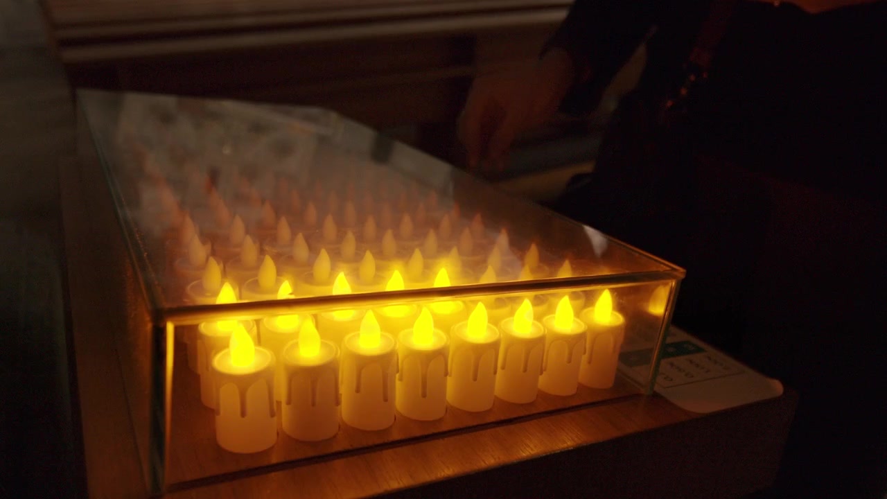 Una persona accende delicatamente candele a batteria in una chiesa, onorando la memoria dei propri cari scomparsi in un momento di quieta riflessione
