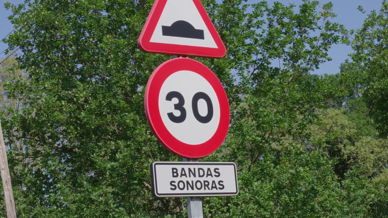 Señales de tráfico claras indican un límite de 30 km/h y bandas sonoras más adelante, bajo la luz brillante del día, con un telón de fondo de paisaje rural verde y frondoso.