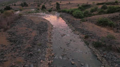 Aerial,-rocky-stream-flows-through-arid-valley-at-dusk-with-purple-clouds