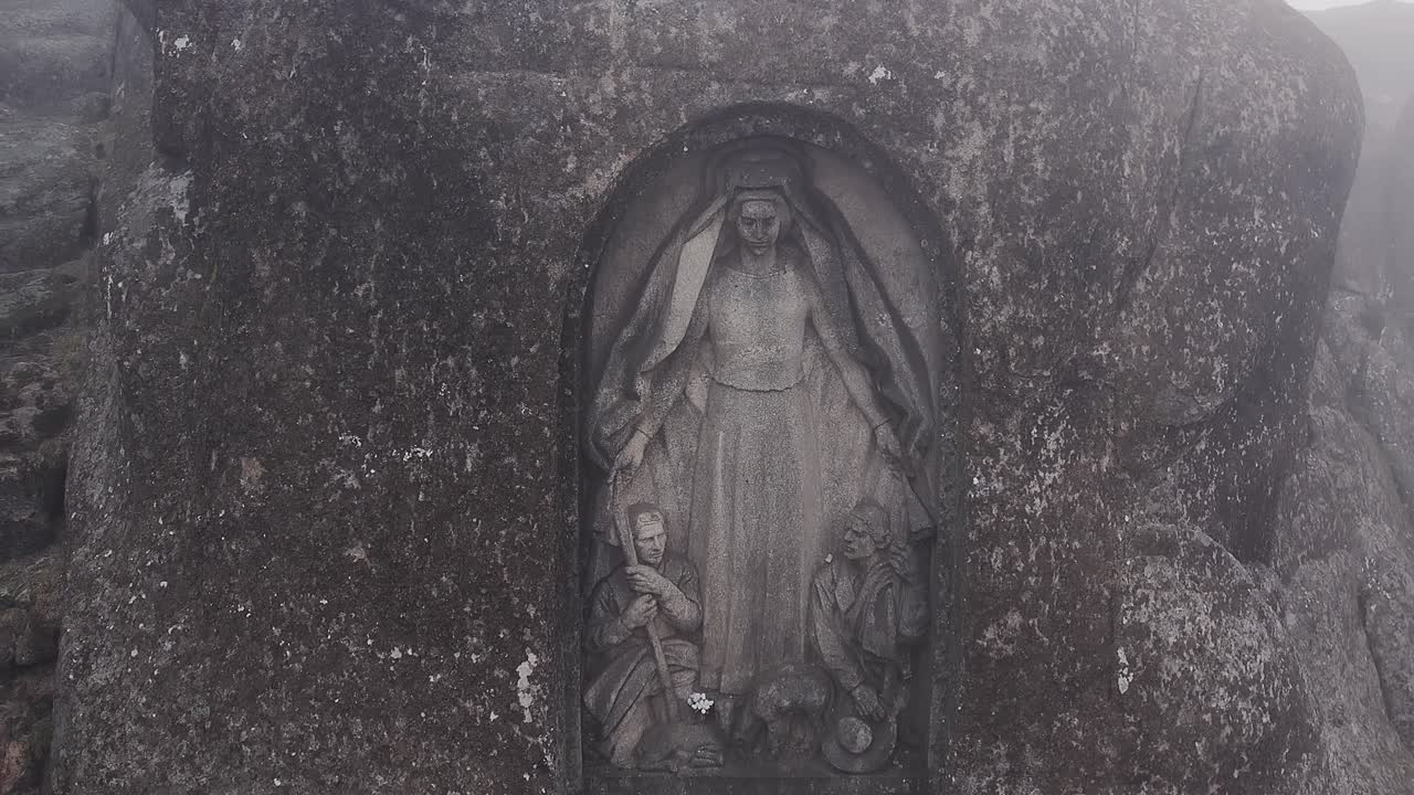 La statua di Nossa Senhora da Boa Estrela, scolpita in una nicchia di granito nella Serra da Estrela, Portogallo, rivela le circostanti formazioni rocciose montane aspre e l'atmosfera nebbiosa mentre il drone si allontana con una veduta aerea.