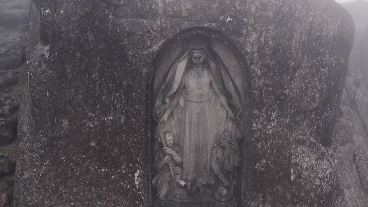 La statua di Nossa Senhora da Boa Estrela, scolpita in una nicchia di granito nella Serra da Estrela, Portogallo, rivela le circostanti formazioni rocciose montane aspre e l'atmosfera nebbiosa mentre il drone si allontana con una veduta aerea.